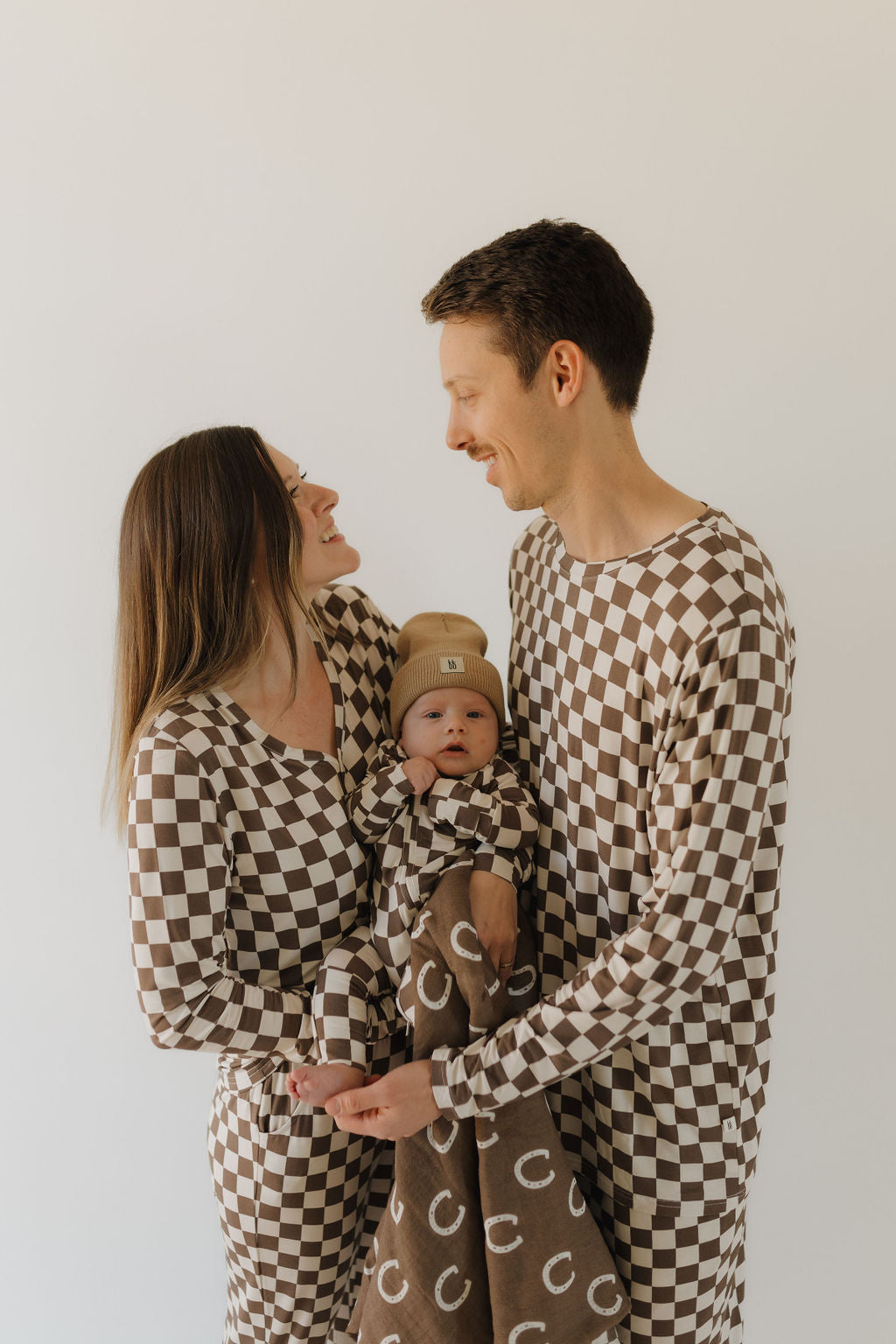 A family of three, wearing forever french baby's matching Bamboo Zip Pajamas in brown and white checkered patterns, stands against a plain background. The woman holds a baby in a breathable fabric blanket with a simple design. Both adults smile at each other.