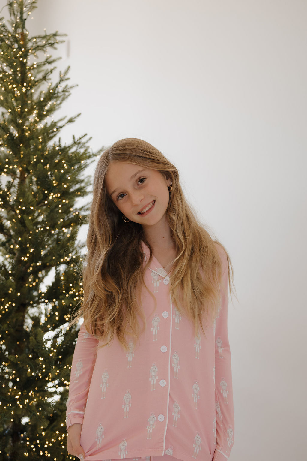 A young girl with long blonde hair smiles beside a decorated Christmas tree with warm white lights, wearing forever french baby's Pre-Teen Flare Bamboo Pajamas | Nutcracker. The background is plain and bright.