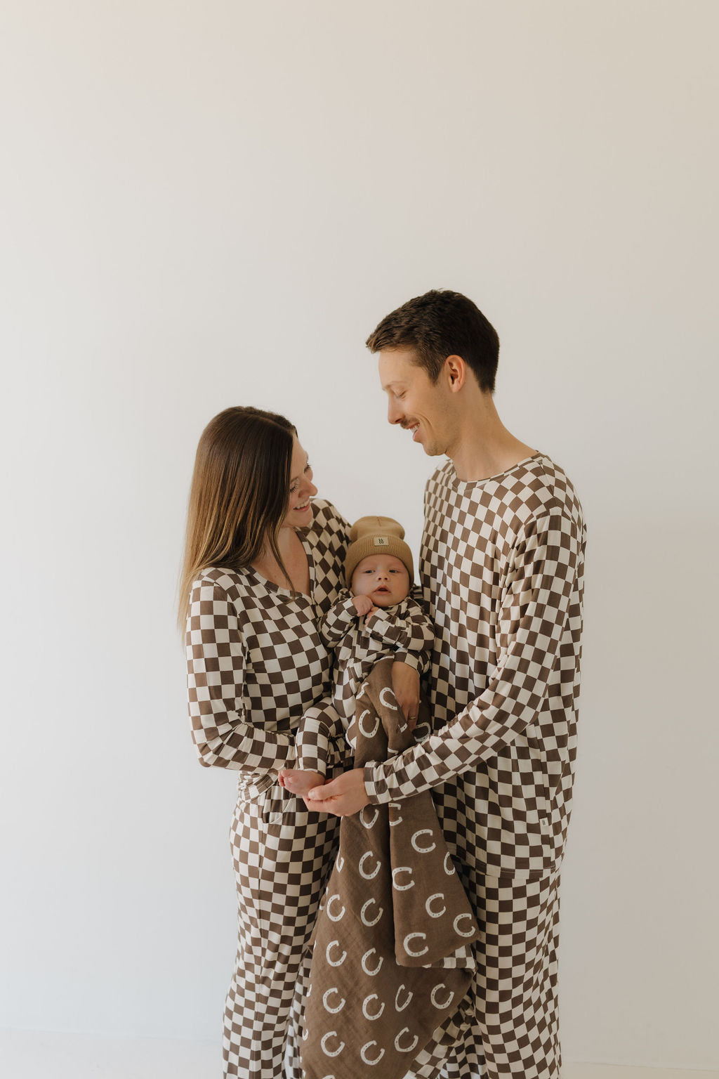 A couple, each cradling a baby in a brown blanket, stands close together against a plain, light background. They exude warmth in matching checkered Bamboo Zip Pajamas by forever french baby, made from breathable fabric.