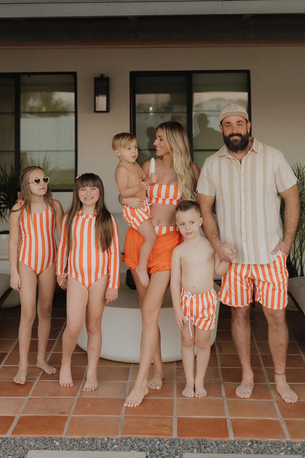 A family of six, dressed in the Poolside Collection's orange and white striped swimwear, stands on a patio. The parents hold a toddler in Forever French Baby attire, alongside three children in Girls Long Sleeve Swimsuits. They smile before a backdrop of modern house exteriors and potted plants.