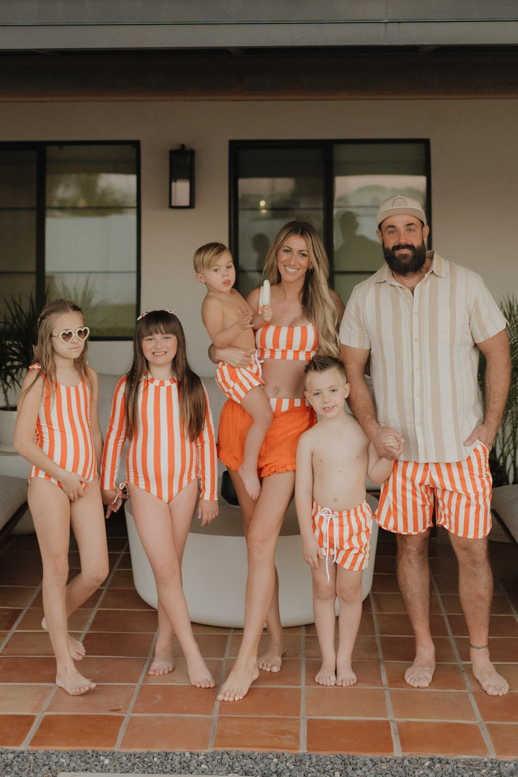 A family of six stands on a patio, all wearing orange and white striped swimwear from the Poolside collection by forever french baby. The mother wears a Women's Two Piece Swimsuit with a high-waisted bikini bottom while holding their youngest child. The other three children smile brightly at the camera.