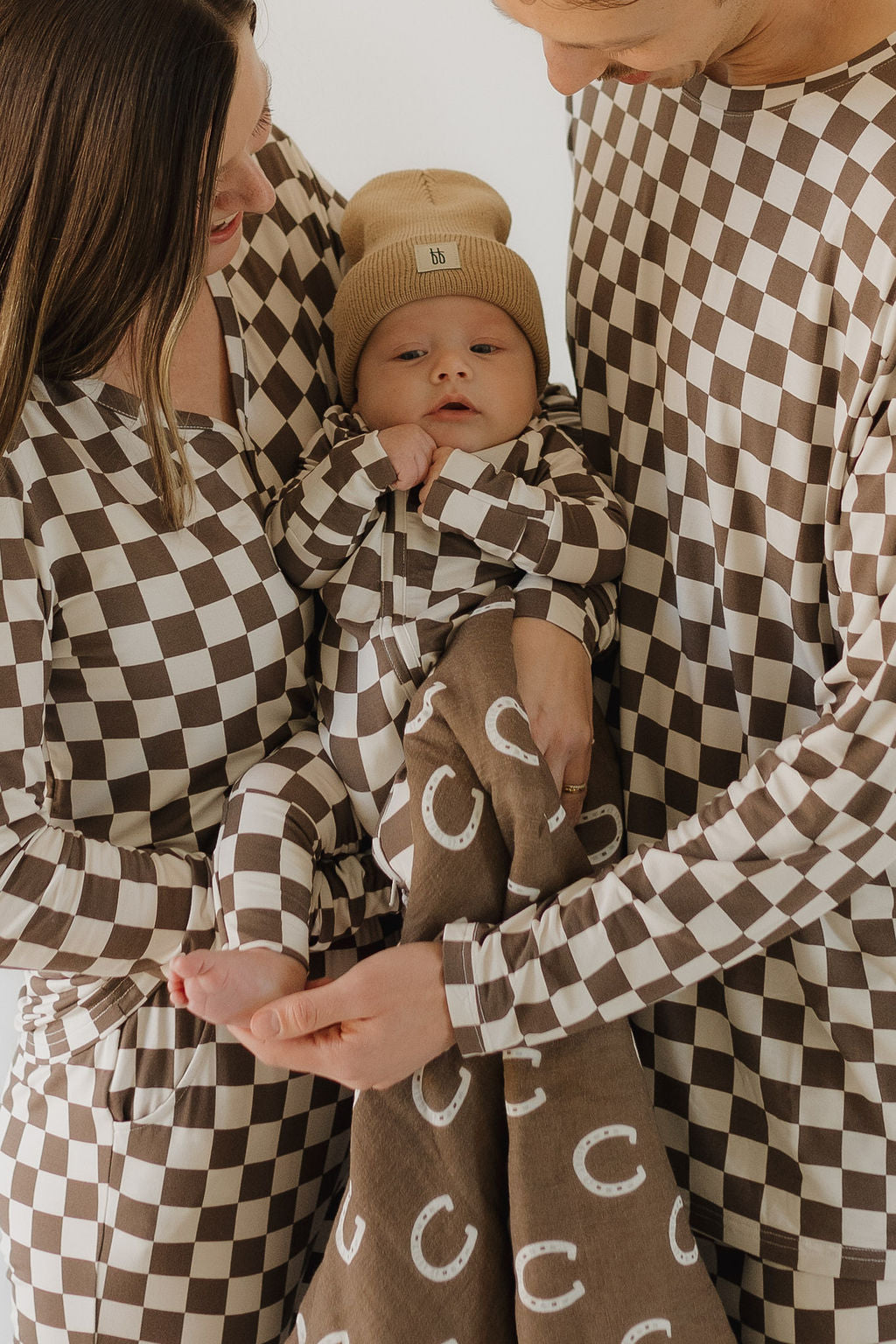 A family of three is wearing matching Bamboo Zip Pajamas in brown and white checkered fabric by forever french baby. The baby, in a beige hat, is held by the parents wrapped in a cozy brown blanket with white "C" patterns. They are close together, smiling warmly.