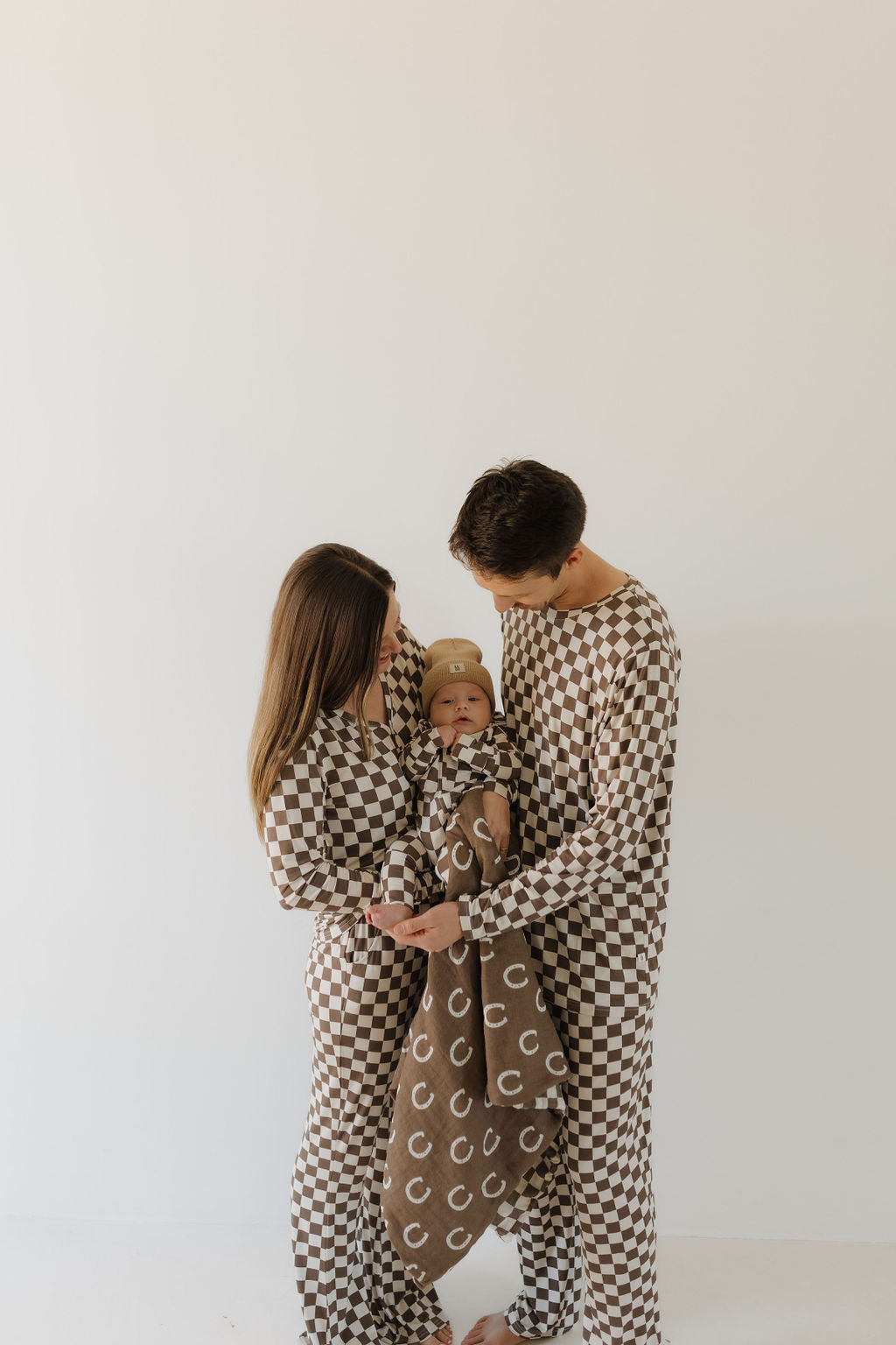 A couple stands holding a baby, all wearing matching brown and white checkered outfits made from breathable fabric. The baby is wrapped in a soft brown blanket with white C's, against a plain white wall. They're wearing Bamboo Zip Pajamas by forever french baby.