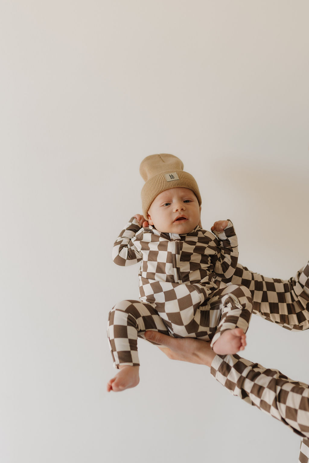 A baby in forever french baby's Bamboo Zip Pajamas | Dutton, featuring a checkered design and breathable fabric, wears a beige knit hat. The infant is held up by adult hands against a plain backdrop, calm and serene, with the image evoking a soft, warm tone.