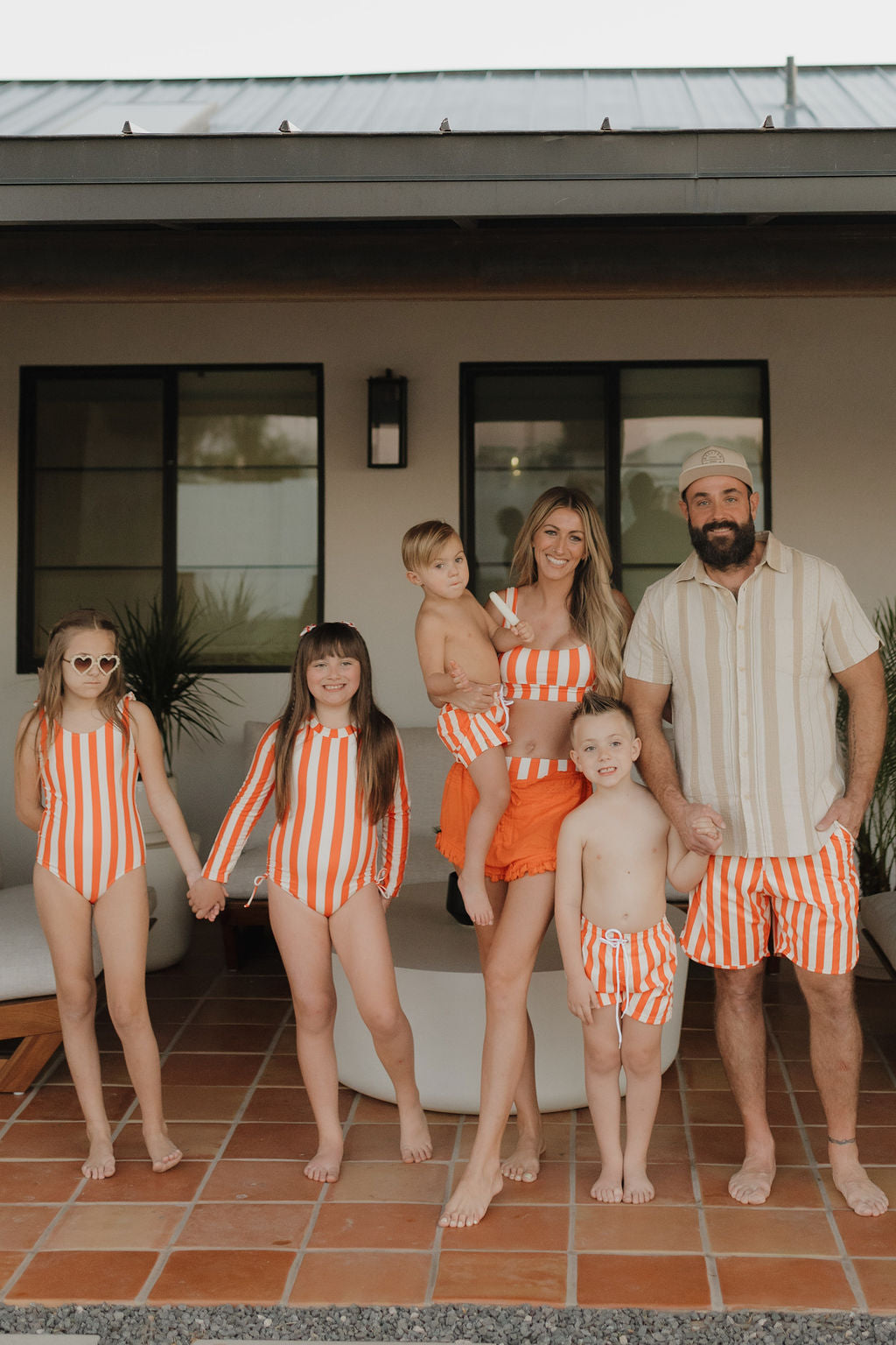 A family of six stands barefoot on a tiled patio, all in matching orange and white swimwear from Forever French Baby's Poolside collection. With the mother cradling a child, their smiles light up against a modern building backdrop with large windows and lush plants.