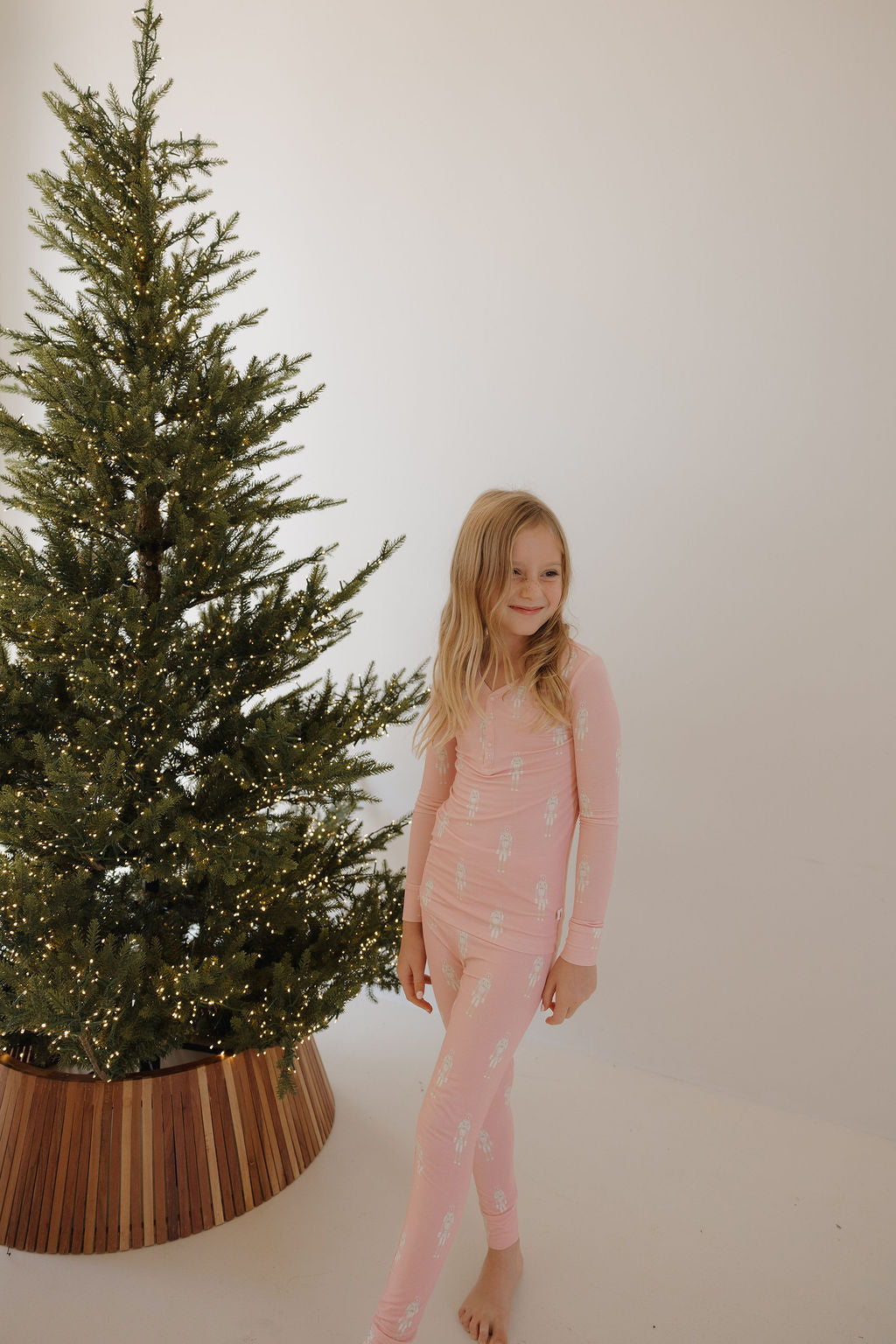 A young girl with long blonde hair stands barefoot near a lit Christmas tree in a minimalist room, wearing forever french baby Bamboo Two Piece Pajamas | Nutcracker in pink.