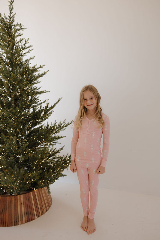 A young girl wears forever french baby's Bamboo Two Piece Pajamas | Nutcracker, featuring a white print, and stands barefoot beside a Christmas tree with white lights. The plain white wall background creates a minimal, festive vibe.