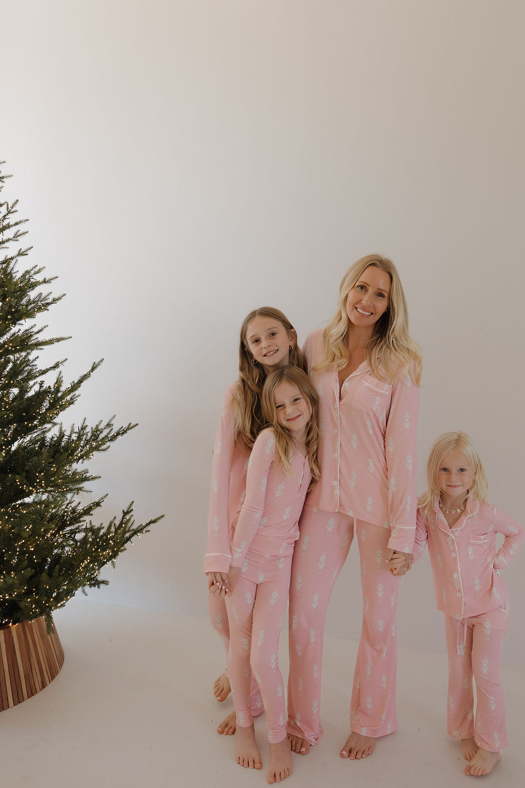 A woman and three young girls smile barefoot by a Christmas tree, all wearing forever french baby Long Sleeve Women's Bamboo Pajamas | Nutcracker, in a bright, minimal room.
