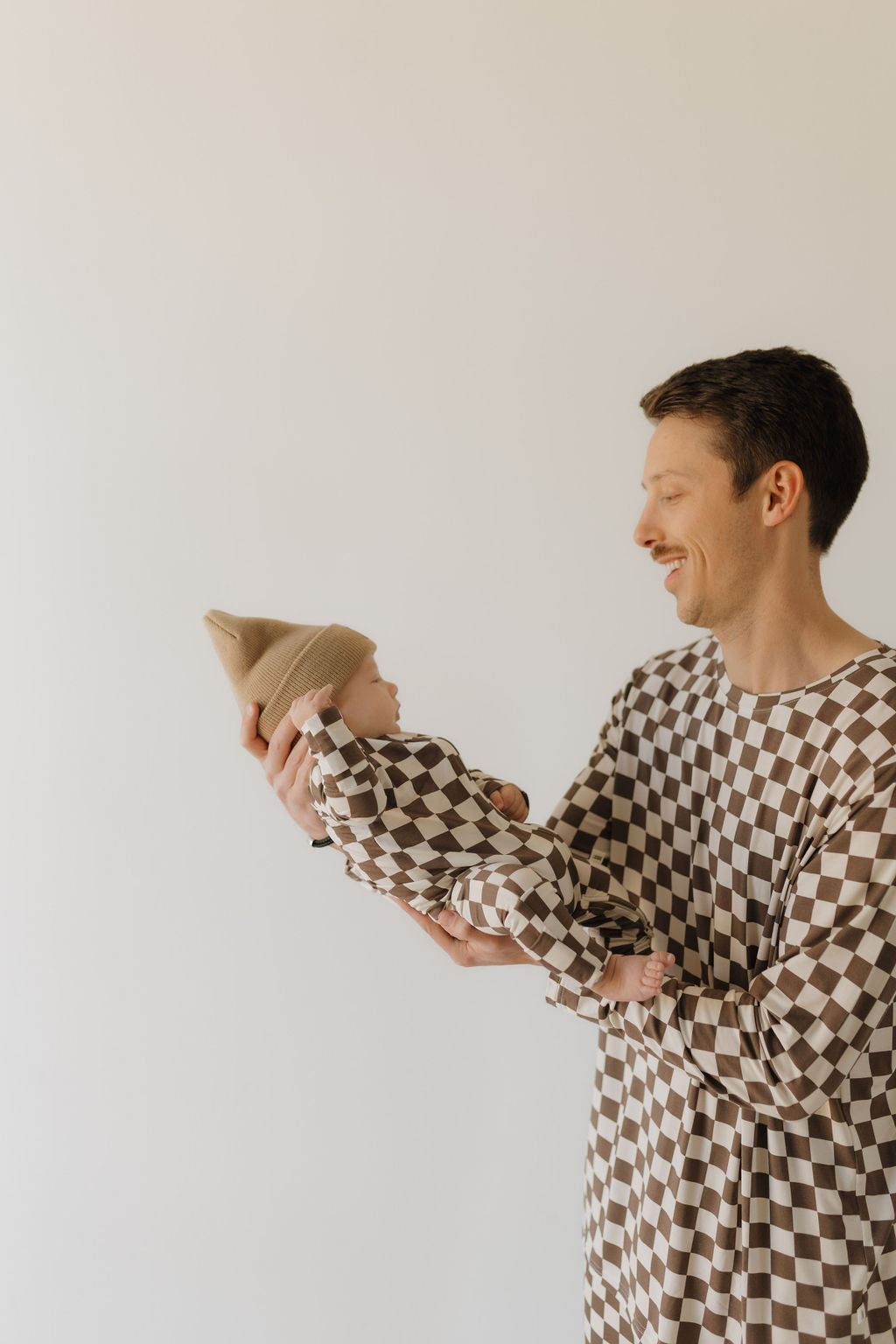 A person holding a baby, both in matching bamboo zip pajamas by forever french baby, featuring brown and white checkered patterns. The baby also wears a tan knitted beanie against an off-white background.