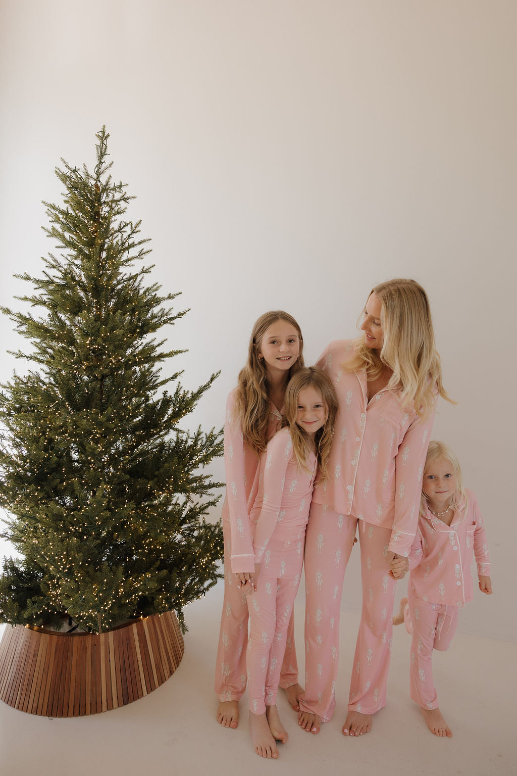 A woman and three young girls, all barefoot and smiling, huddle together by a glowing Christmas tree. They wear matching forever french baby Bamboo Two Piece Pajamas | Nutcracker, radiating warmth and holiday joy against a light background.