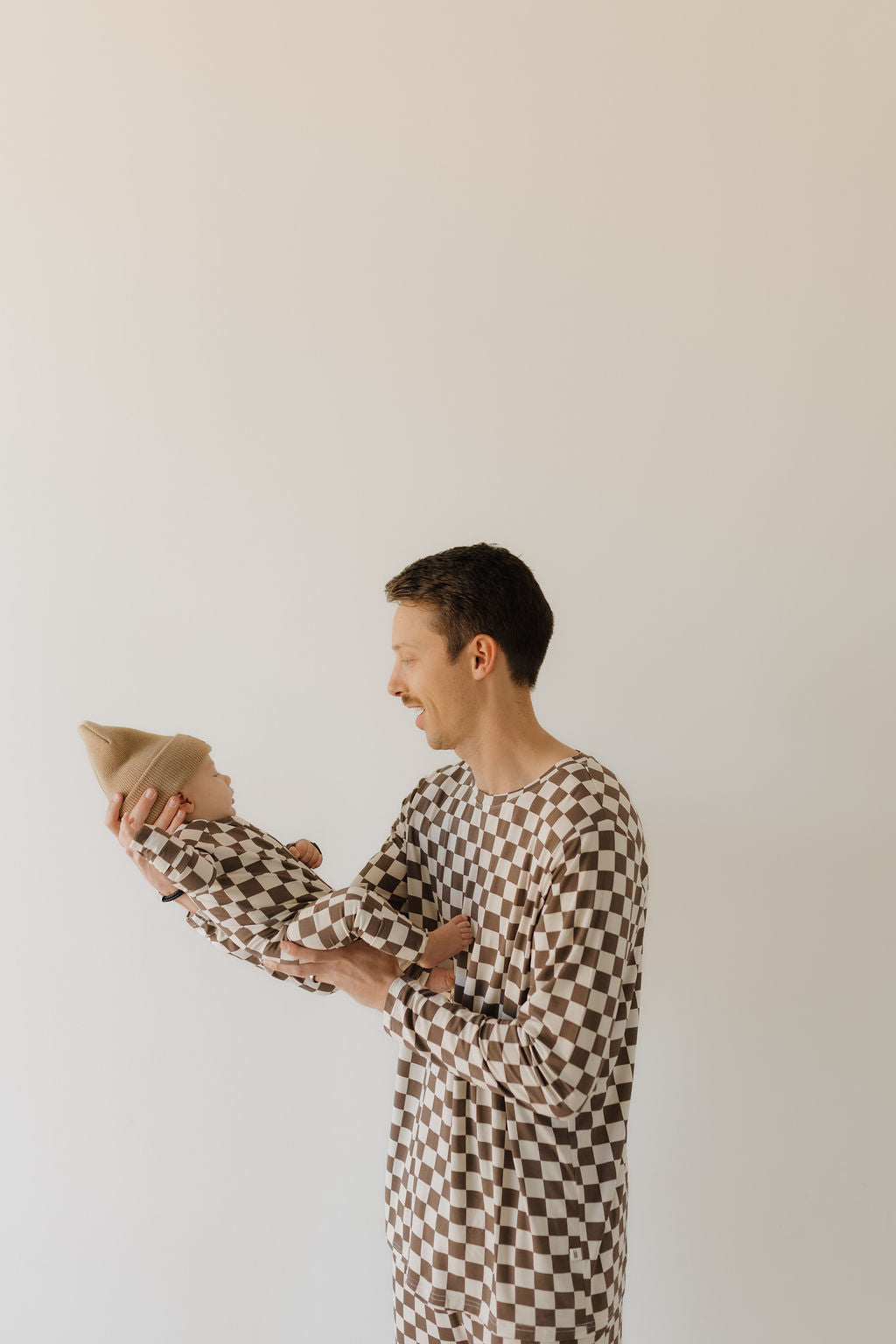 A man holds a baby, both wearing matching "Bamboo Zip Pajamas | Dutton" by forever french baby. The baby's beige hat and their checkered outfits made from breathable fabric are highlighted against a plain light background as the man smiles warmly at the baby.