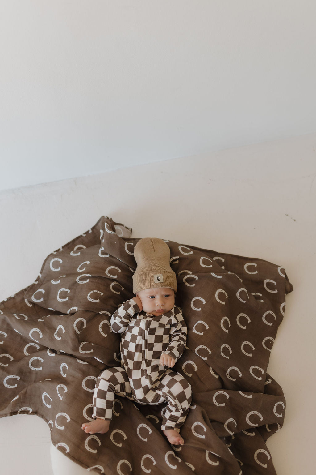 A baby in forever french baby Bamboo Zip Pajamas lies on a brown blanket adorned with white crescent moons, wearing a beige hat and brown checkered outfit. The hypo-allergenic, breathable fabric adds comfort on the light-colored surface.