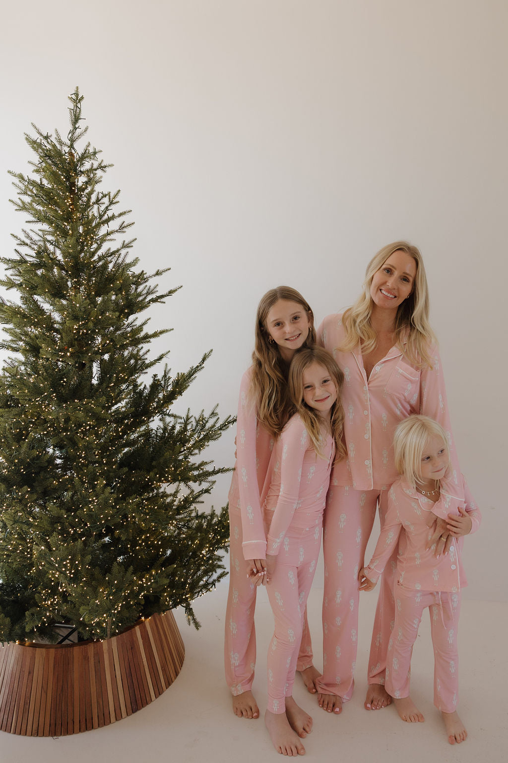 A woman and three girls smile by a festive tree, all wearing forever french baby’s Pre-Teen Flare Bamboo Pajamas | Nutcracker—cute, matching pink holiday sets that are cozy, hypoallergenic, and perfect for family moments.