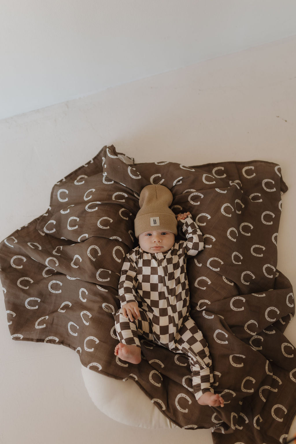 A baby rests on a round brown blanket with white crescents, wearing a cozy knit cap and hypo-allergenic clothing. The serene scene is completed with Bamboo Zip Pajamas by forever french baby, as the little one looks up, hand near head.