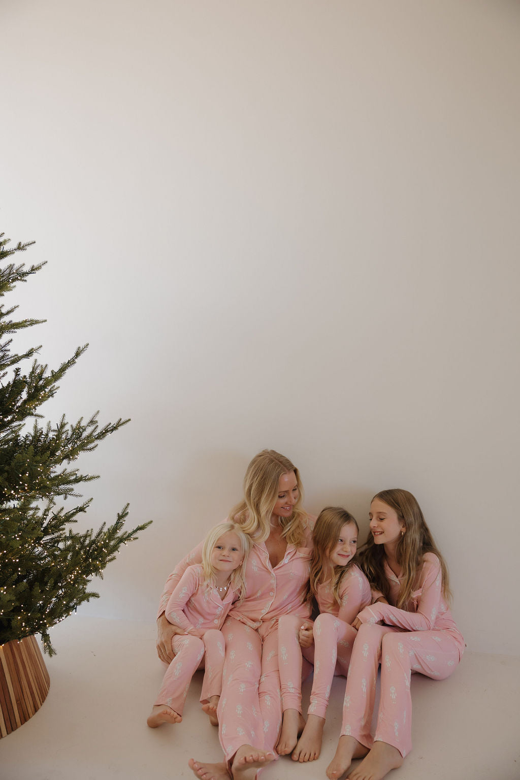 A woman and three girls, smiling together by a Christmas tree, wear matching Pre-Teen Flare Bamboo Pajamas | Nutcracker from forever french baby—cozy, hypo-allergenic lounge sets made for comfort and holiday cheer.