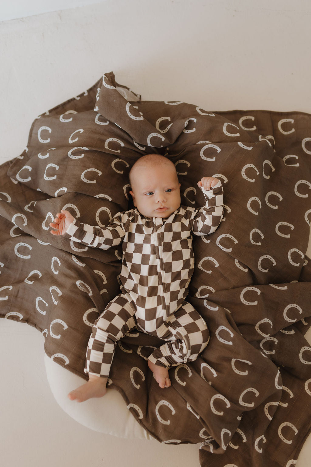 A baby is comfortably lying on a soft surface, donning the "Bamboo Zip Pajamas | Dutton" by forever french baby in a checkered brown and white design. Wrapped in a cozy brown blanket with white circles, they gaze upwards, enjoying the breathable fabric.