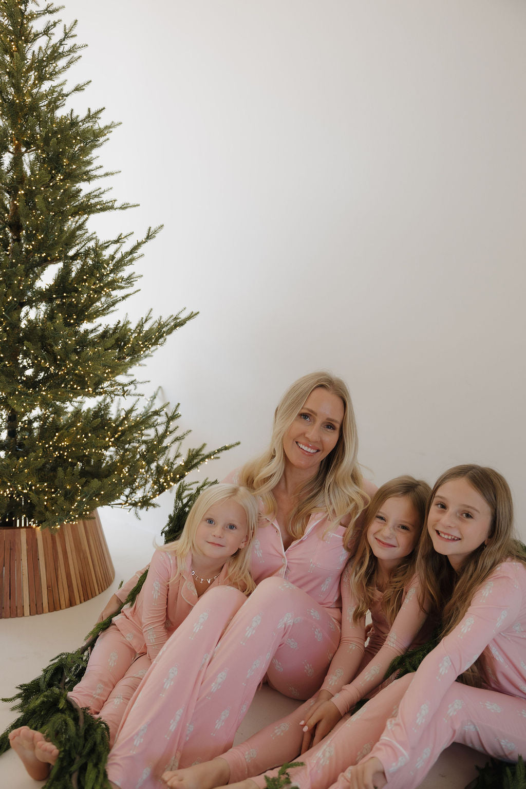 A woman and three young girls, smiling in matching forever french baby Long Sleeve Women's Bamboo Pajamas | Nutcracker, sit by a Christmas tree with white lights for a festive, cozy holiday photo.