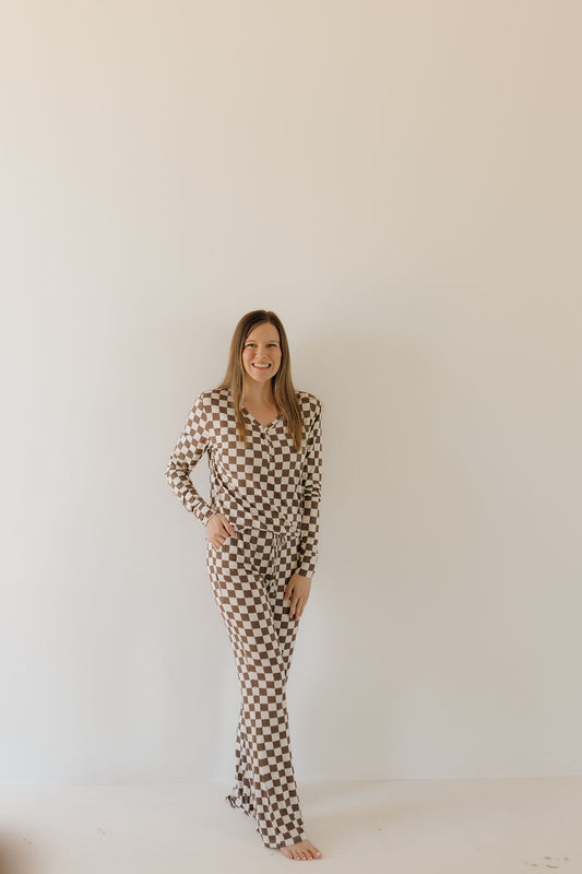 A person stands against a plain light-colored wall, wearing the Women's Bamboo Pajama in brown and white checks by forever french baby. Made from breathable bamboo fabric, they have long hair, are barefoot, and smile with one hand on their hip.