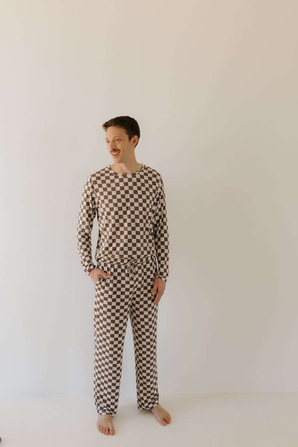 A person poses against a plain background wearing a "Men's Bamboo Pajama | Dutton" by forever french baby. The hypo-allergenic, brown and white checkerboard patterned long-sleeved top and pants, crafted from soft bamboo viscose, are perfect for comfort. Hands in pockets, barefoot, they gaze to the side.