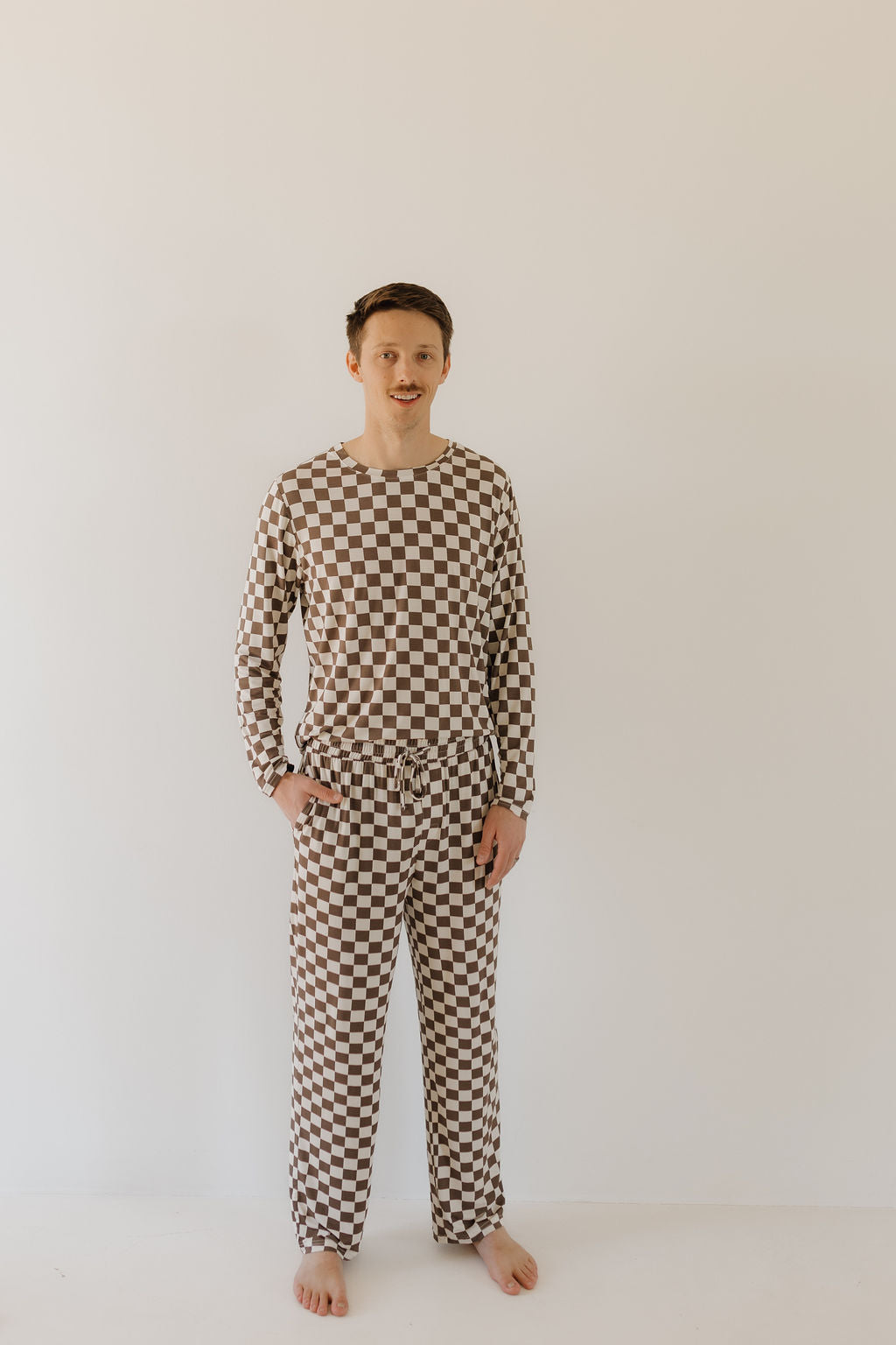 Barefoot, a person models the Men's Bamboo Pajama | Dutton by forever french baby, featuring a checkered brown and white pattern against a plain white wall. With hands in pockets, this breathable ensemble epitomizes cozy charm while the wearer gazes confidently at the camera.