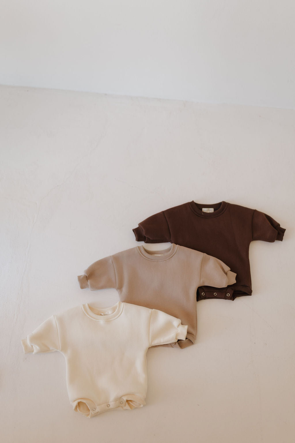 Three neutral-toned Forever French Baby 'Sweatshirt Romper | Driftwood' onesies—cream, beige, and brown—are displayed on a light surface. They each feature long sleeves and convenient side snap buttons for easy diaper changes.