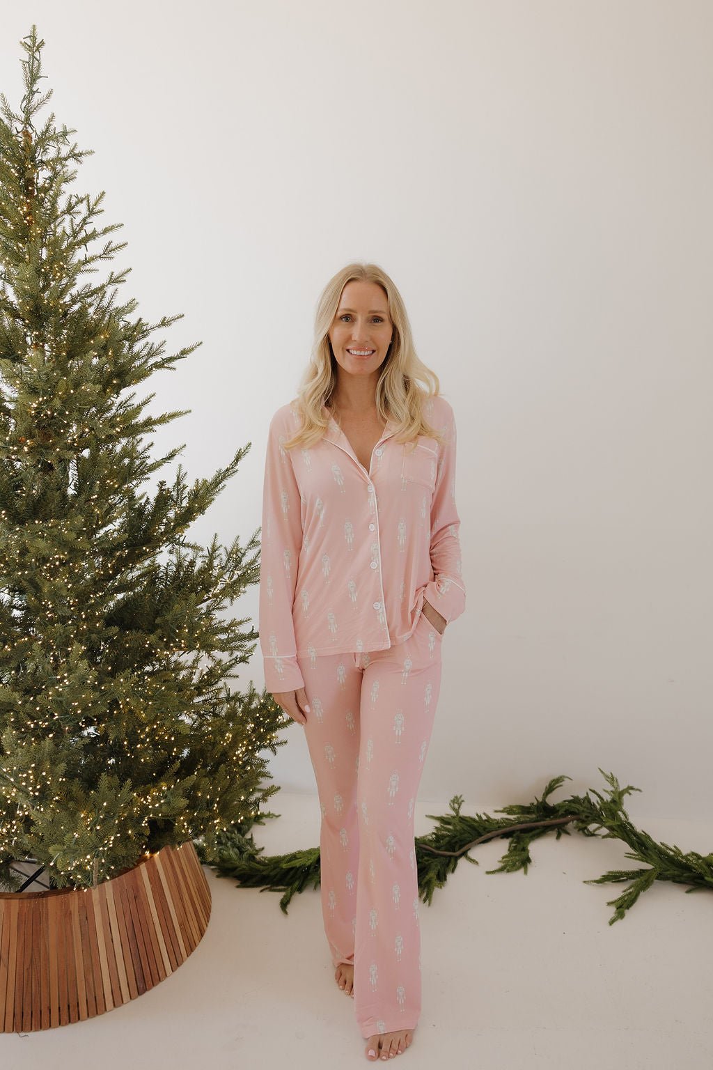 A woman with long blonde hair, barefoot, stands by a Christmas tree in forever french baby Long Sleeve Women's Bamboo Pajamas | Nutcracker—pink with a white pattern. Green garland decorates the white floor nearby.