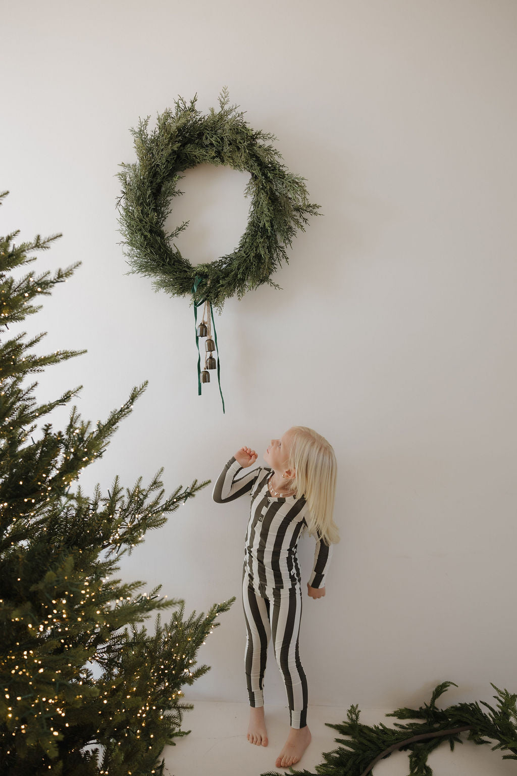 A young child wearing forever french baby Bamboo Two Piece Pajamas in Fireside Stripe gazes up at a green wreath with bells on a white wall, with a lit and decorated Christmas tree visible on the left.
