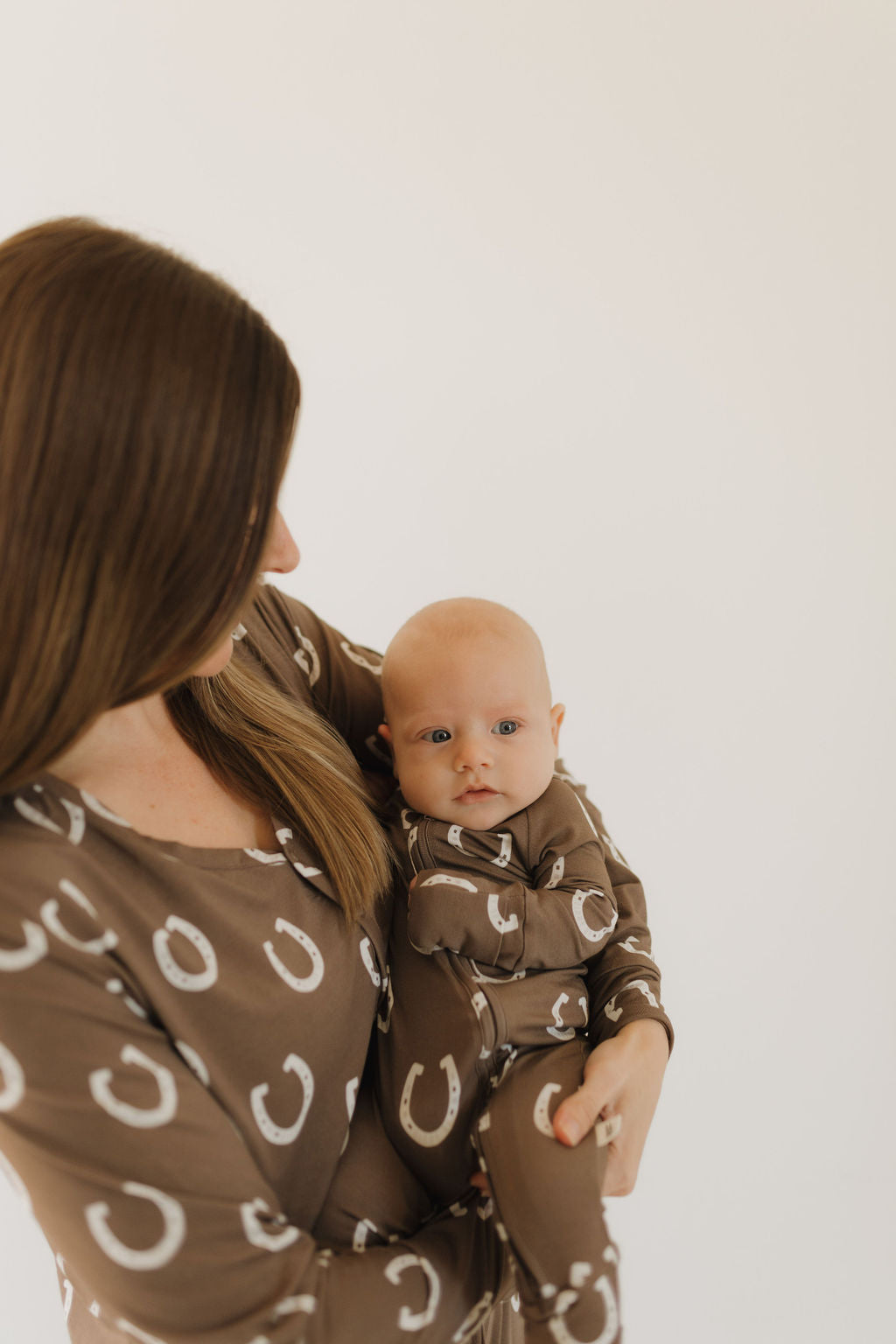 A woman with long brown hair holds a baby wearing forever french baby's matching Bamboo Zip Pajamas | Giddy Up in brown with a white horseshoe pattern. The breathable, hypo-allergenic clothing complements the plain, light-colored background as the baby gazes at the camera.