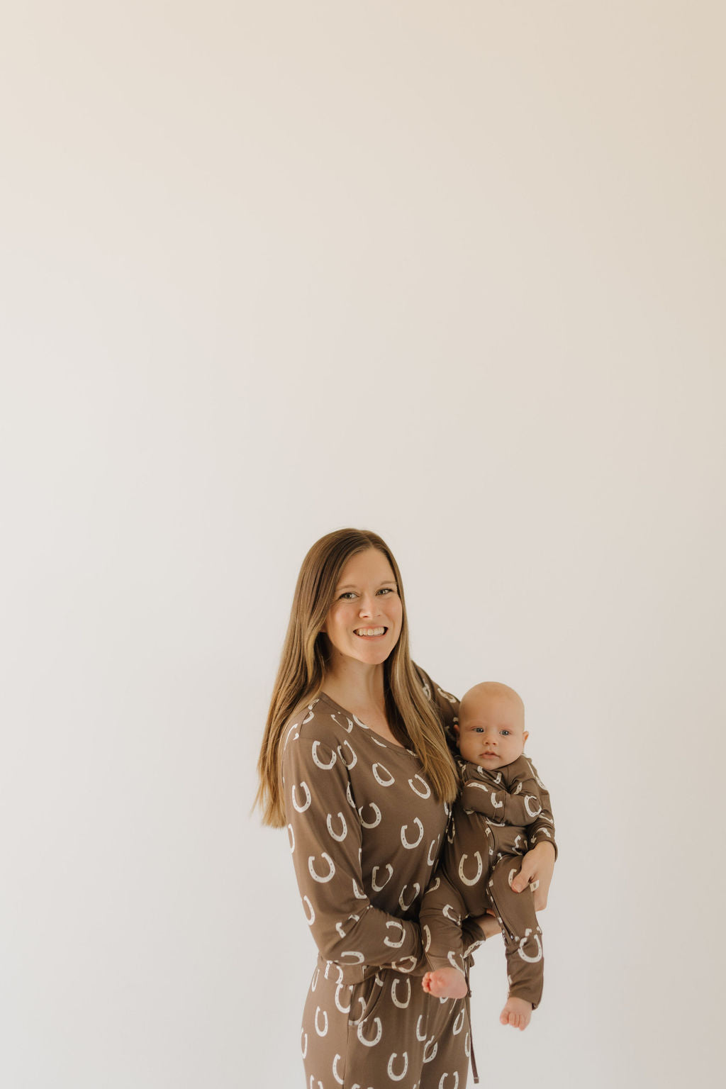 A woman with long brown hair in a breathable outfit with white horseshoe patterns from forever french baby's Bamboo Zip Pajamas | Giddy Up collection holds a baby in matching attire against a light background.