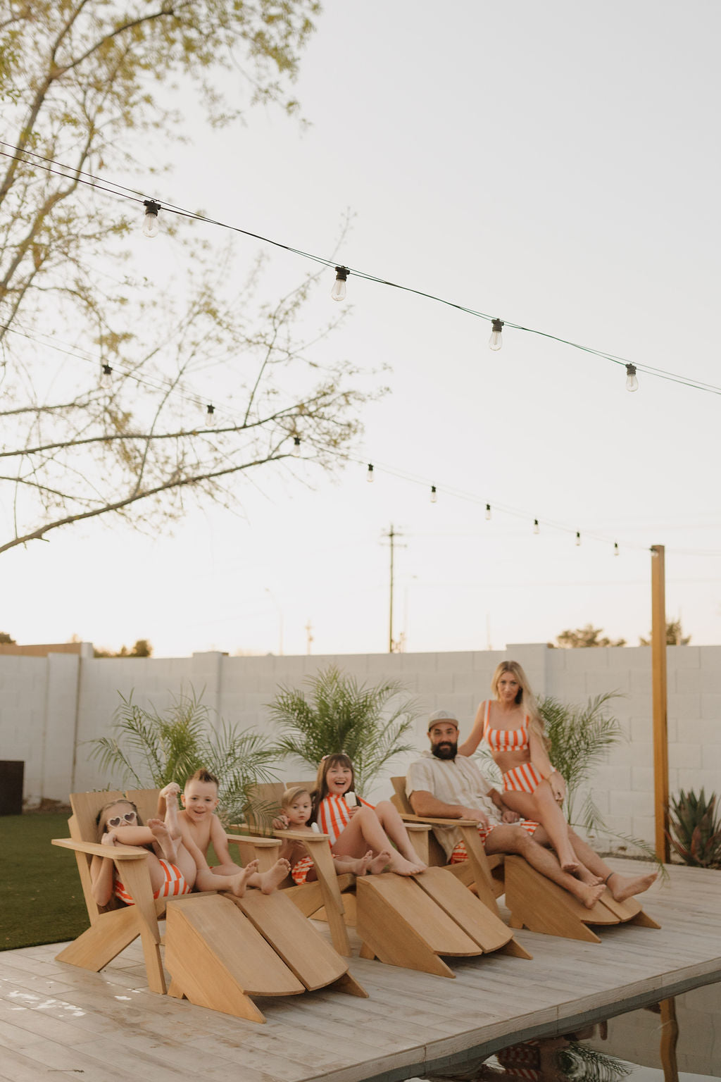 A family of six relaxes by the pool on wooden loungers, all wearing forever french baby Men's Boardshorts from the Poolside Collection in matching orange and white stripes. String lights twinkle above, while lush plants frame the scene against a clear sky.