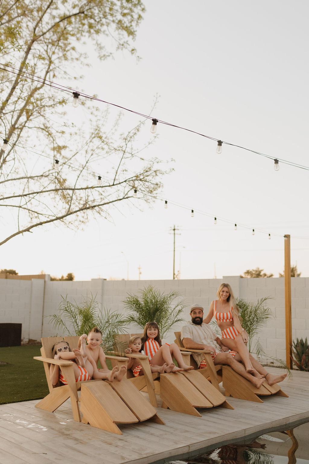 A family of five relaxes on wooden lounge chairs by the pool, wearing matching attire from the Poolside collection of forever french baby. String lights and potted plants under a clear sky enhance the serene ambiance.