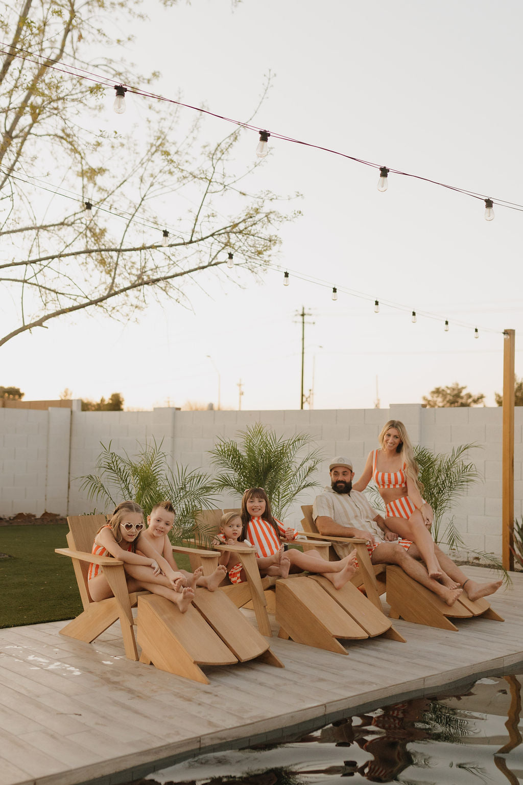 A family of six relaxes by the pool in wooden chairs, wearing vibrant orange and white striped Child Boardshorts from forever french baby. Trees and string lights create a charming backdrop for the sunny, relaxed scene.