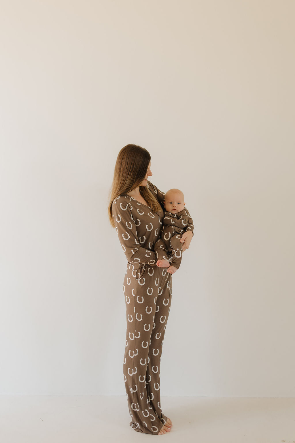 A woman with long brown hair stands against a plain backdrop, barefoot and gazing lovingly at a baby. Both wear forever french baby's Bamboo Zip Pajamas in the Giddy Up design, featuring breathable and hypo-allergenic fabric with white horseshoe patterns.