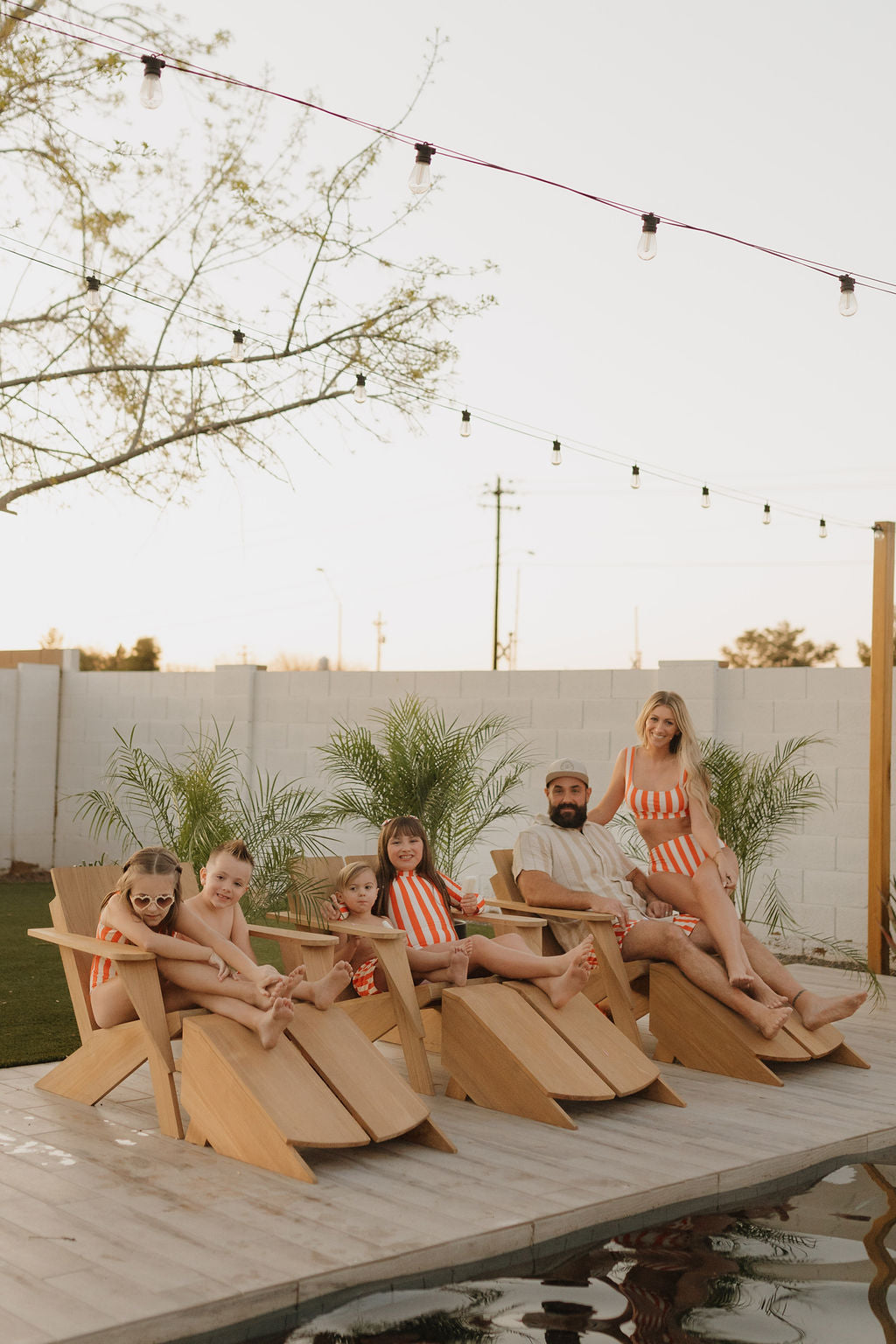 A family of six lounges by the pool on wooden chairs. The children joyfully play in boardshorts and matching red and white striped swimsuits from forever french baby's Poolside collection, while adults relax casually amid twinkling string lights and lush potted plants.