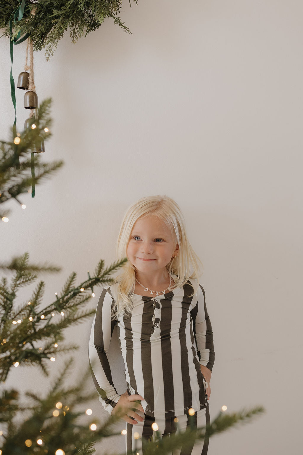 A young blonde girl smiles by a decorated Christmas tree, wearing forever french baby Bamboo Two Piece Pajamas in Fireside Stripe, with festive greenery and bells hanging above her.