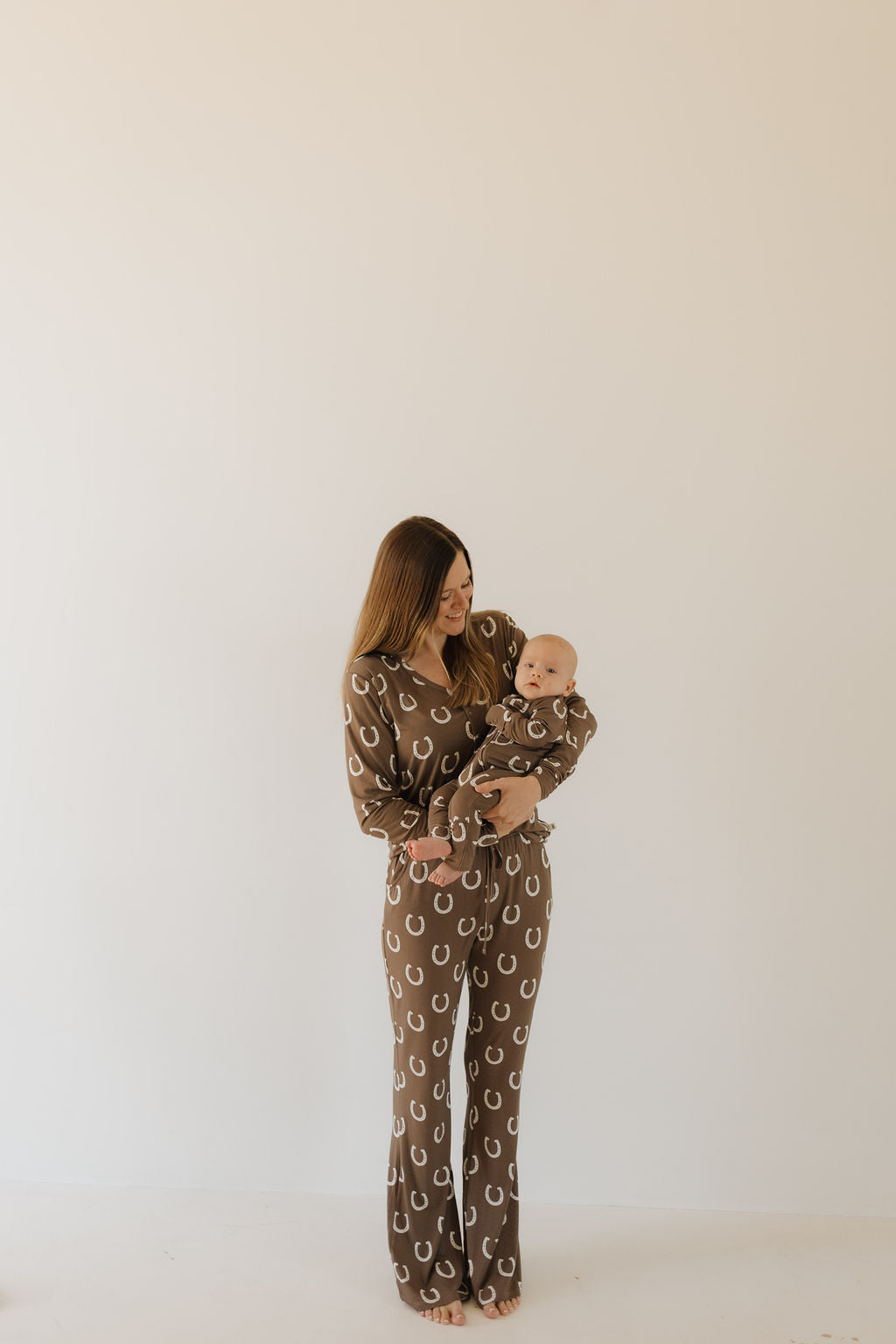 A woman in "Giddy Up" bamboo zip pajamas by forever french baby, featuring a circular pattern, holds a baby wearing the same hypo-allergenic set. They stand against a plain light background, gazing at each other.