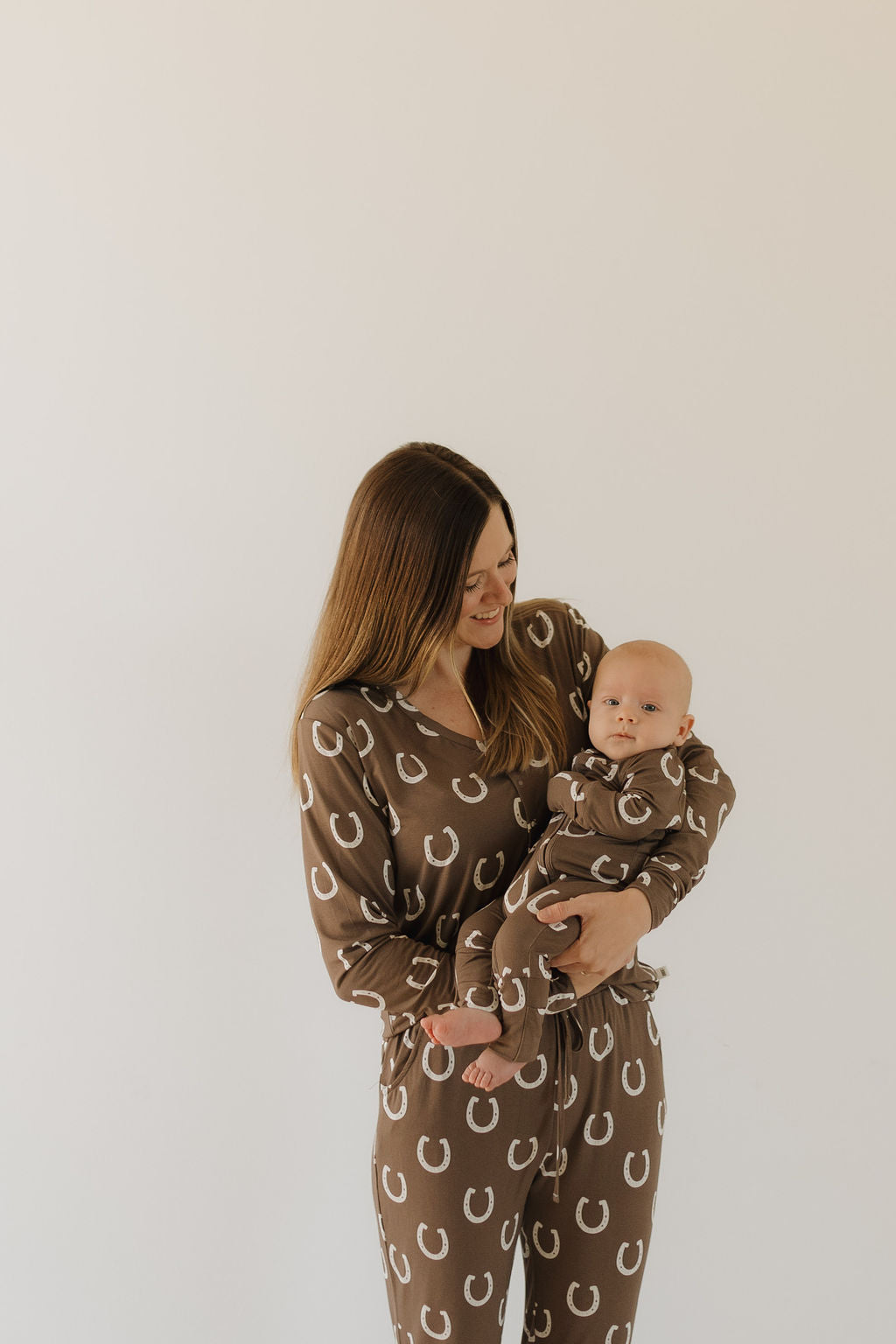 A woman cradles a baby, both in matching forever french baby Bamboo Zip Pajamas | Giddy Up, featuring brown fabric with white horseshoe patterns. Against a plain white background, she smiles at the baby.