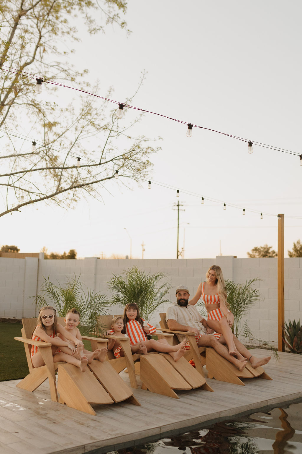 A family lounges on wooden chairs by the pool in matching orange and white striped swimsuits from forever french baby's Poolside Collection. A child wears a Girls Long Sleeve Swimsuit, while string lights twinkle above and potted plants form a lush backdrop.