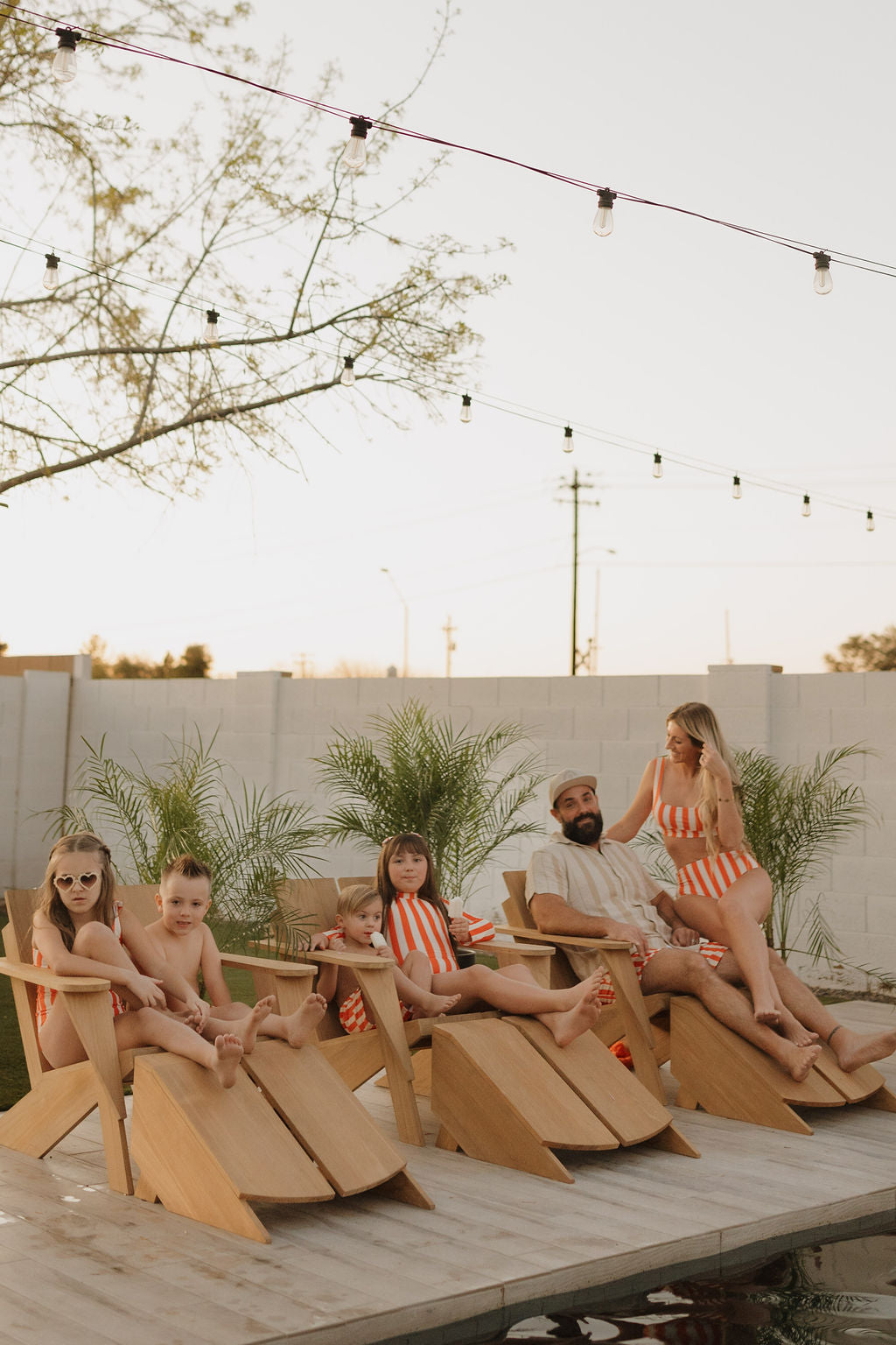 A family of six relaxes on wooden lounge chairs by the pool, showcasing the vibrant Poolside Collection from forever French baby. They wear matching red and white striped Girls Sleeveless Swimsuit under a clear sky, with palm plants and string lights enhancing the cheerful atmosphere.