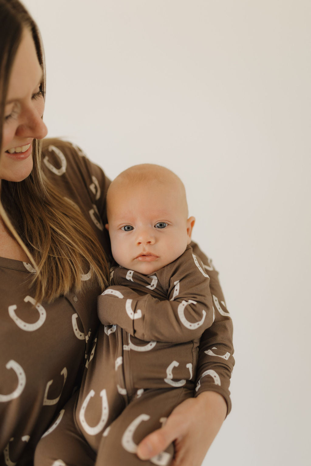 A woman and a baby wear matching "Bamboo Zip Pajamas | Giddy Up" from forever french baby, featuring a brown fabric with white horseshoe patterns. The breathable attire complements the baby's direct gaze at the camera as the woman smiles lovingly in front of a soft neutral background.