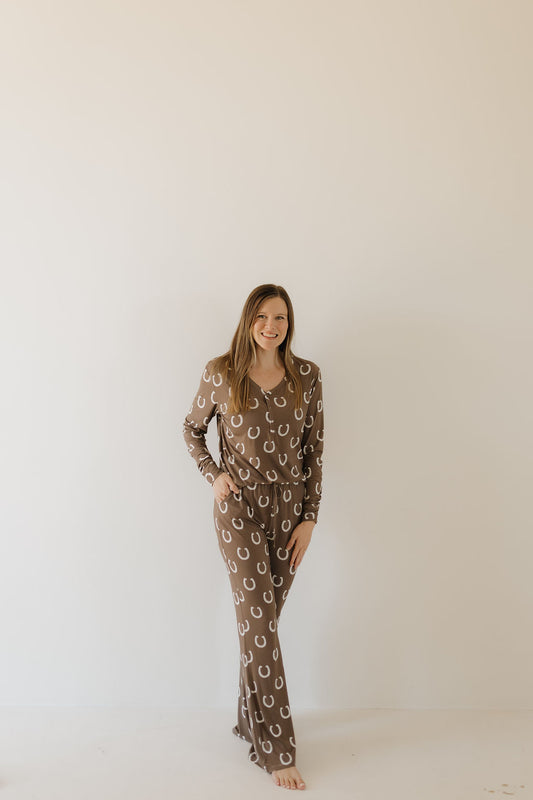 A person stands barefoot against a light wall, wearing the Women's Bamboo Pajama | Giddy Up from forever french baby. Featuring a horseshoe pattern and breathable fabric, these pajamas offer comfort and style. Smiling with one hand in their pocket, they exude relaxed confidence.