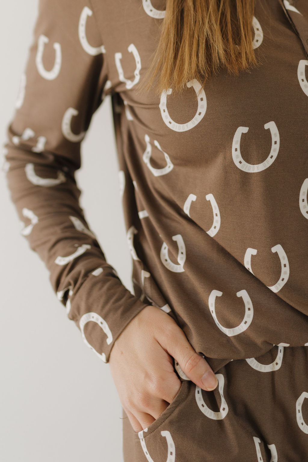 A person in the Women's Bamboo Pajama - Giddy Up by forever french baby dons a brown outfit with a white horseshoe pattern. Made from breathable fabric, the long-sleeve top and bottoms ensure comfort as the hand rests casually in the pocket, partially revealing their hair.