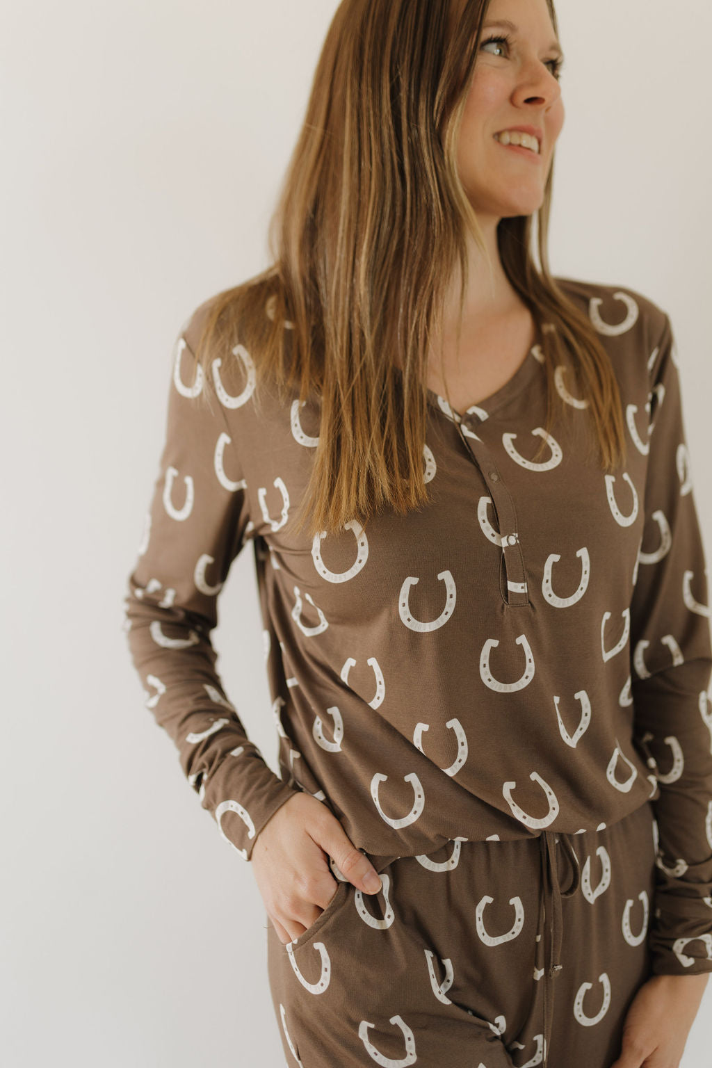 A woman with long brown hair smiles while looking to the side, wearing forever french baby's Women's Bamboo Pajama, "Giddy Up," with a white horseshoe pattern against a plain white background. The breathable fabric ensures comfort and style.