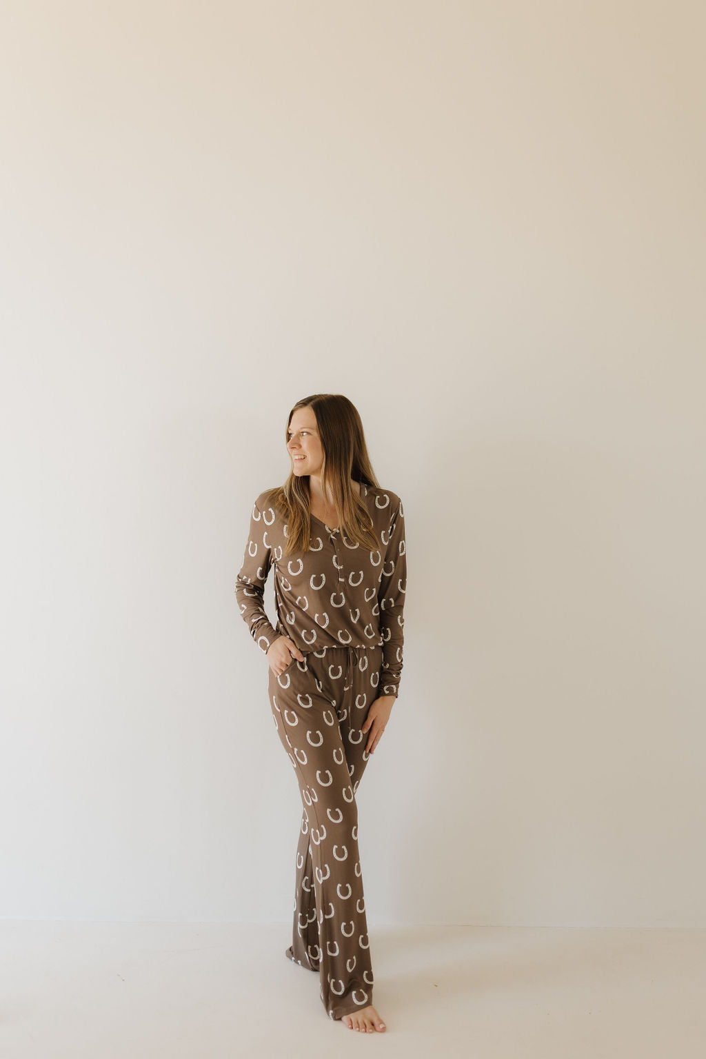 A woman stands against a plain wall, wearing the "Women's Bamboo Pajama | Giddy Up" by forever french baby. The brown outfit features a white horseshoe pattern and is made from breathable fabric. She has one hand in her pocket and is smiling slightly as she looks to the side.