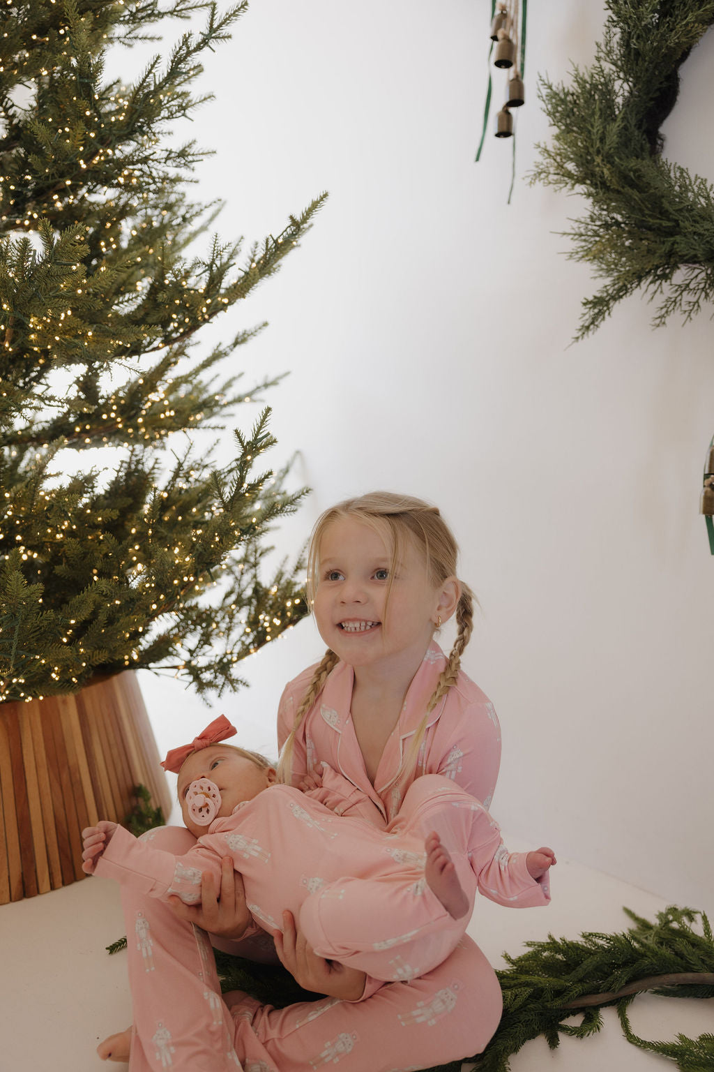 A young girl in pink pajamas smiles, holding a baby wearing forever french baby's Long Sleeve Toddler Bamboo Pajamas | Nutcracker by a Christmas tree and wreaths.