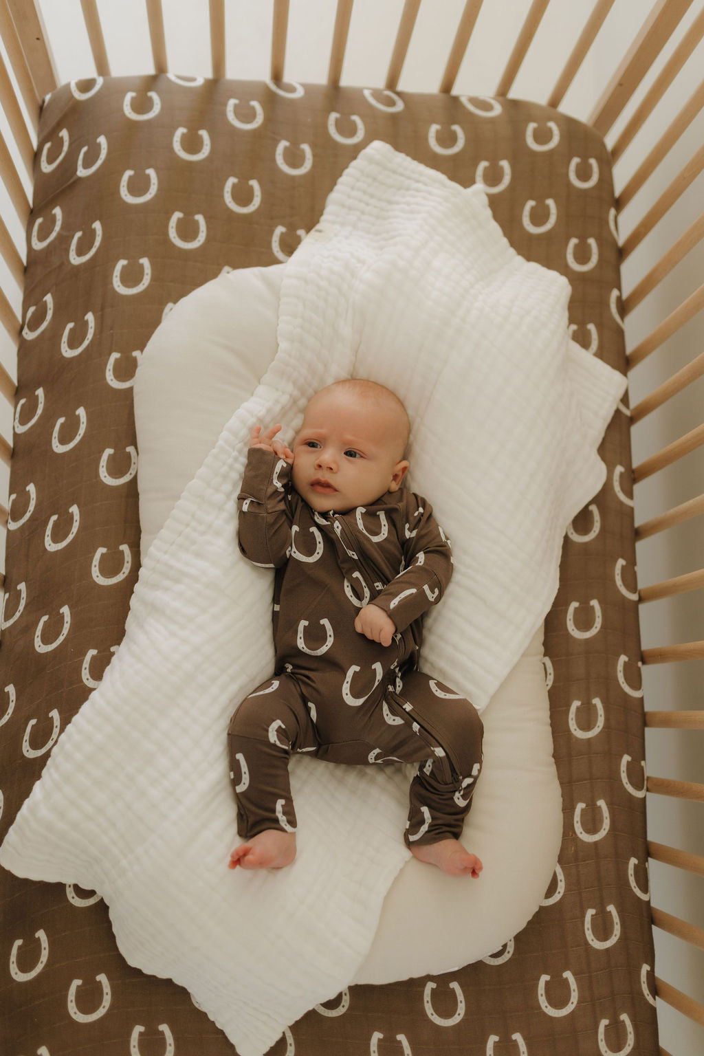 A baby lies in a crib on a brown sheet with white horseshoe patterns, wearing Bamboo Zip Pajamas | Giddy Up by forever french baby. The child rests on a white pillow and blanket, while the wooden slat crib ensures comfort and style with its breathable fabric ensemble.