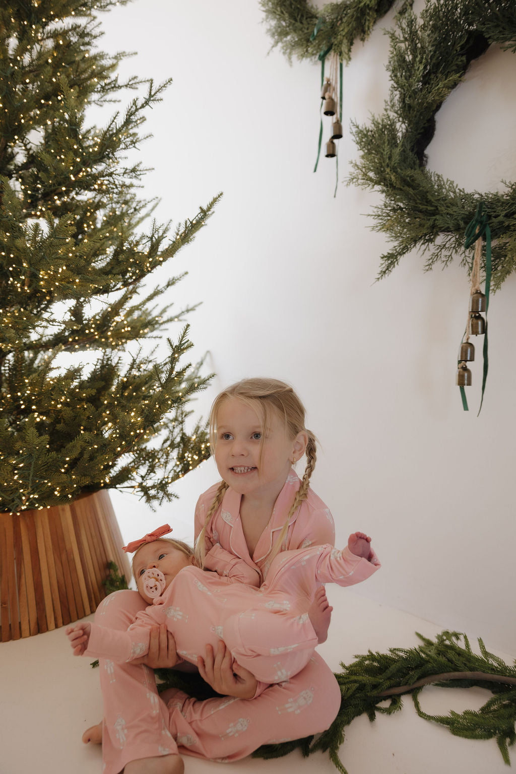 A young girl in forever french baby Bamboo Zip Pajamas | Nutcracker holds a baby, also in breathable bamboo sleepwear, as they smile together on the floor by a Christmas tree and festive wreaths.