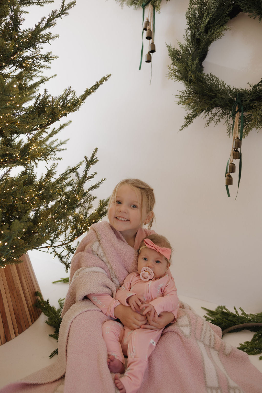 Two young children sit on the floor by a Christmas tree and wreath. The older one smiles at the camera while holding a baby in forever french baby's Bamboo Zip Pajamas | Nutcracker, with a pacifier. Both are wrapped in a pink blanket.