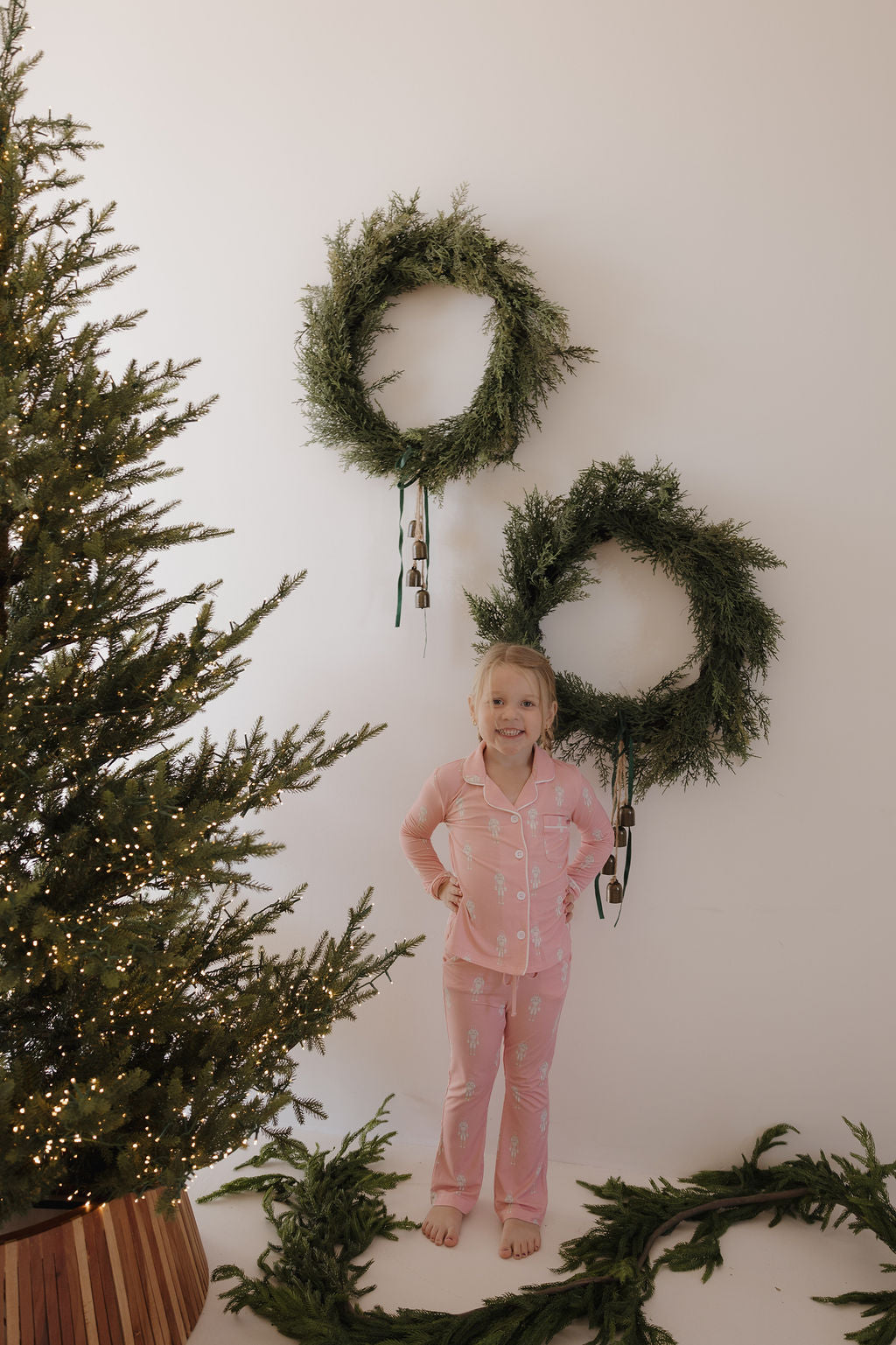 A young child in forever french baby Long Sleeve Toddler Bamboo Pajamas | Nutcracker stands barefoot before a white wall with two green wreaths. A lit Christmas tree and pine garland add to the festive, cozy scene.