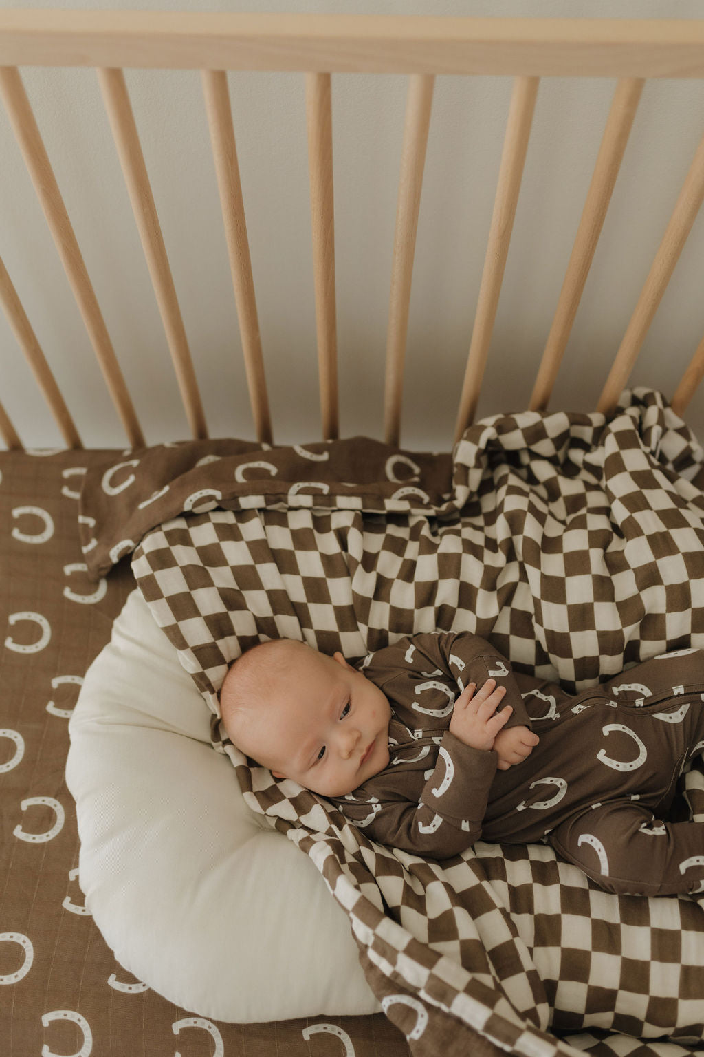 A baby rests in a wooden crib with cozy bedding, wearing Bamboo Zip Pajamas by forever french baby featuring crescent patterns. They're wrapped in a matching checkered blanket, while the crib sheet displays similar brown and white crescent designs for all-around comfort.